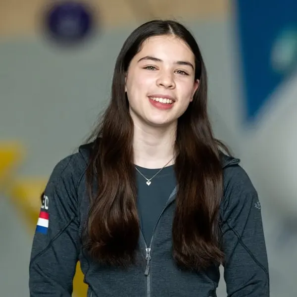 Een jonge vrouw met lang donker haar, gekleed in een donker jasje met een patch in de vorm van de Nederlandse vlag, glimlacht naar de camera tegen een wazige achtergrond binnenshuis.